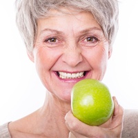 Smiling older woman eating green apple with all-on-4 dental implant denture