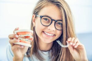 Girl with glasses holding model teeth with braces in one hand and clear aligner in the other